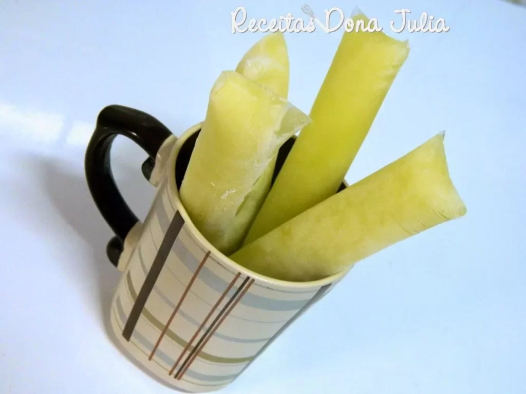 Vários geladinhos de milho verde em saquinhos transparentes, com uma cor amarela vibrante e cremosa, dispostos sobre uma mesa com algumas espigas de milho ao fundo.