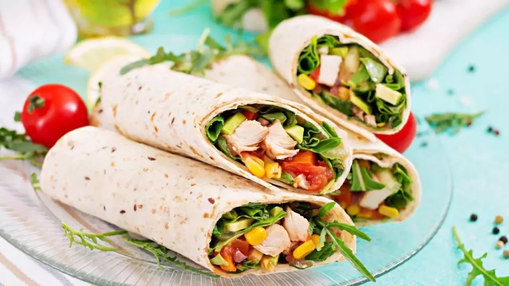 "Um wrap de tortilla integral cortado ao meio na diagonal, revelando camadas coloridas de frango desfiado, fatias verdes de abacate, tomate vermelho, cebola roxa e folhas de alface fresca."
