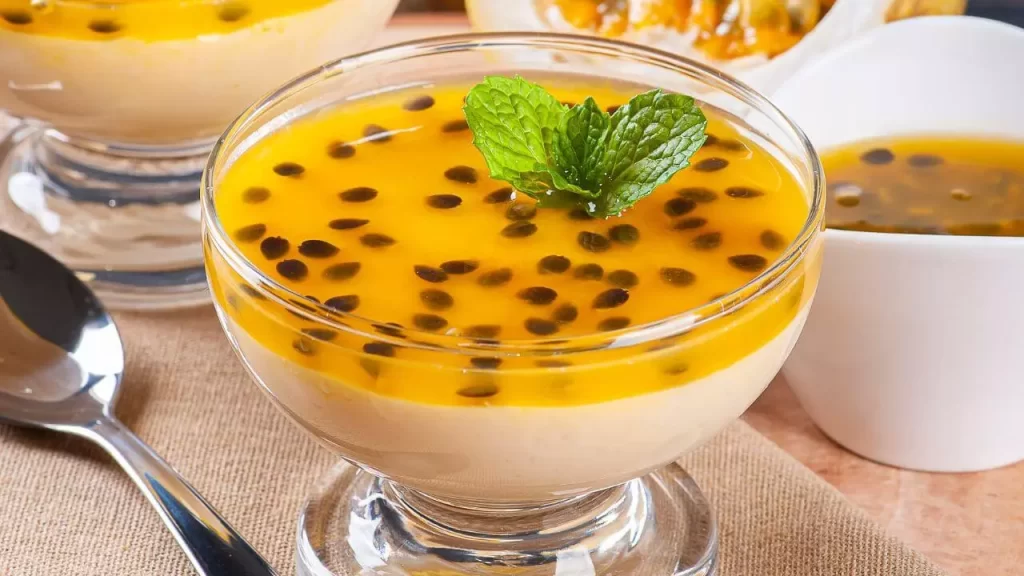 "Uma travessa de vidro com mousse de maracujá amarelo claro e aerado, coberto por uma geleia brilhante com sementes de maracujá, decorada com um raminho de hortelã."