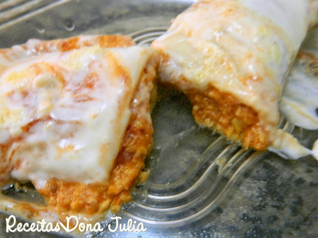 "Panqueca de frango" "Uma travessa refratária com panquecas enroladas, cobertas por um molho de tomate vibrante e uma camada de queijo gratinado dourado com pedacinhos de milho aparecendo no recheio."