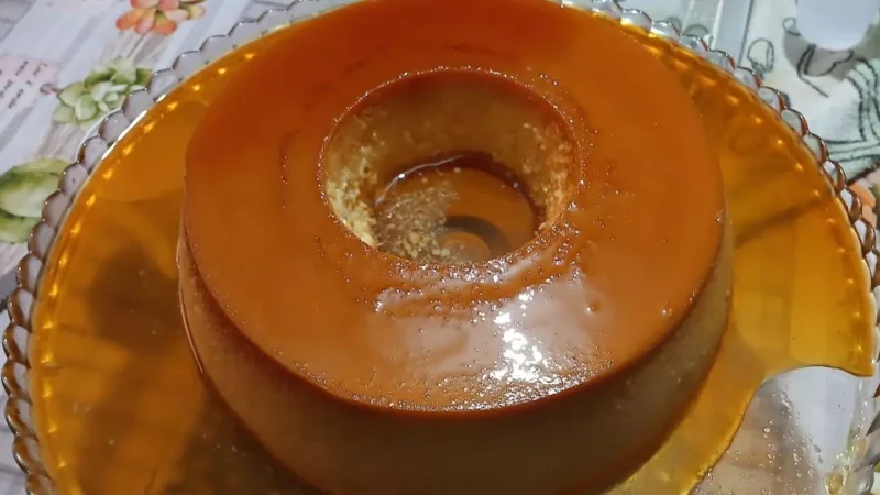 "Um pudim de pão alto e dourado em um prato de servir, com uma calda de caramelo escura e brilhante escorrendo pelas laterais e pedacinhos sutis de uvas-passas aparecendo no miolo úmido."